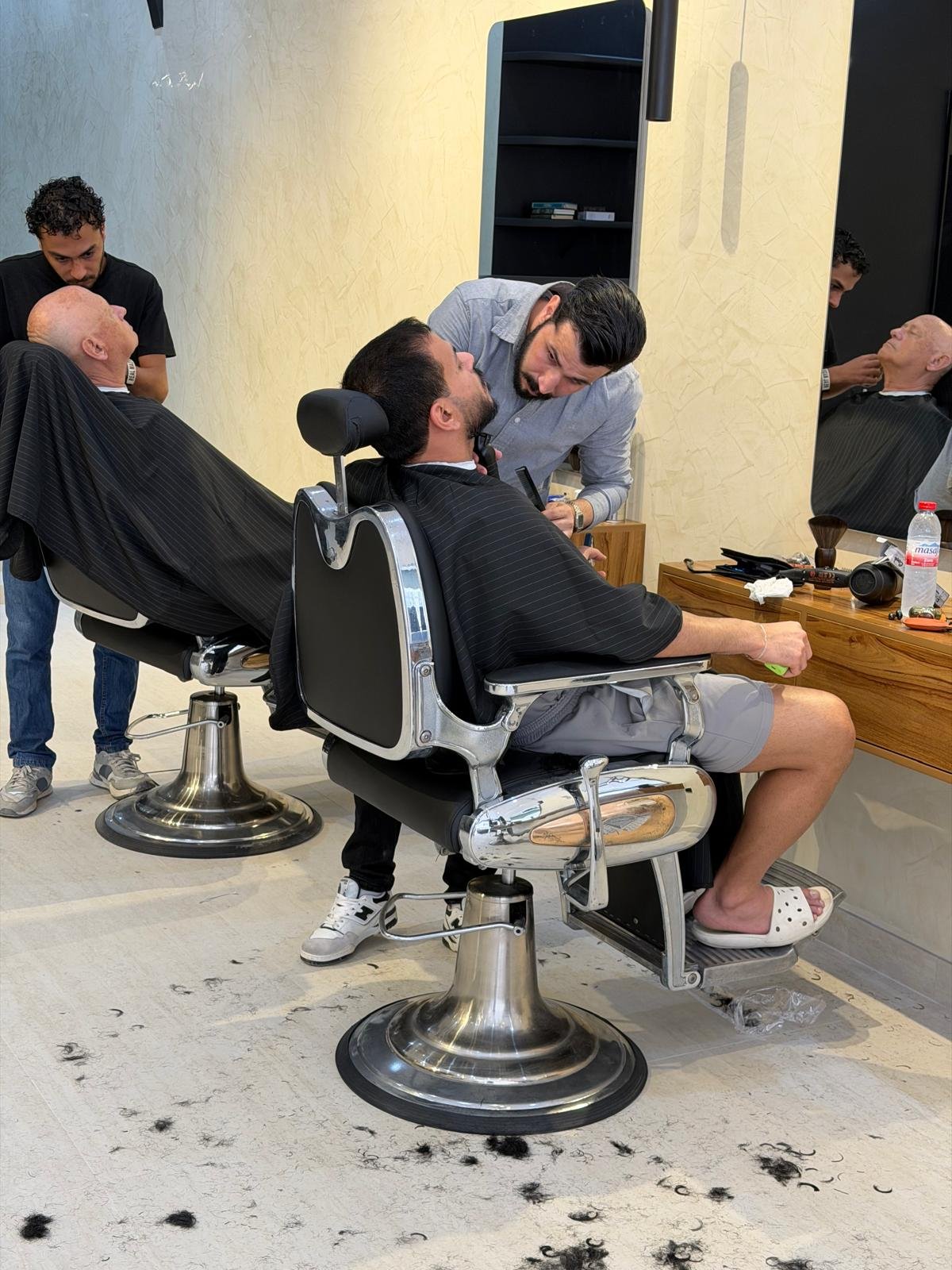Barber trimming a reclined client's beard with clippers while a second barber works the next chair at Brass Barber in Al Barsha, Dubai