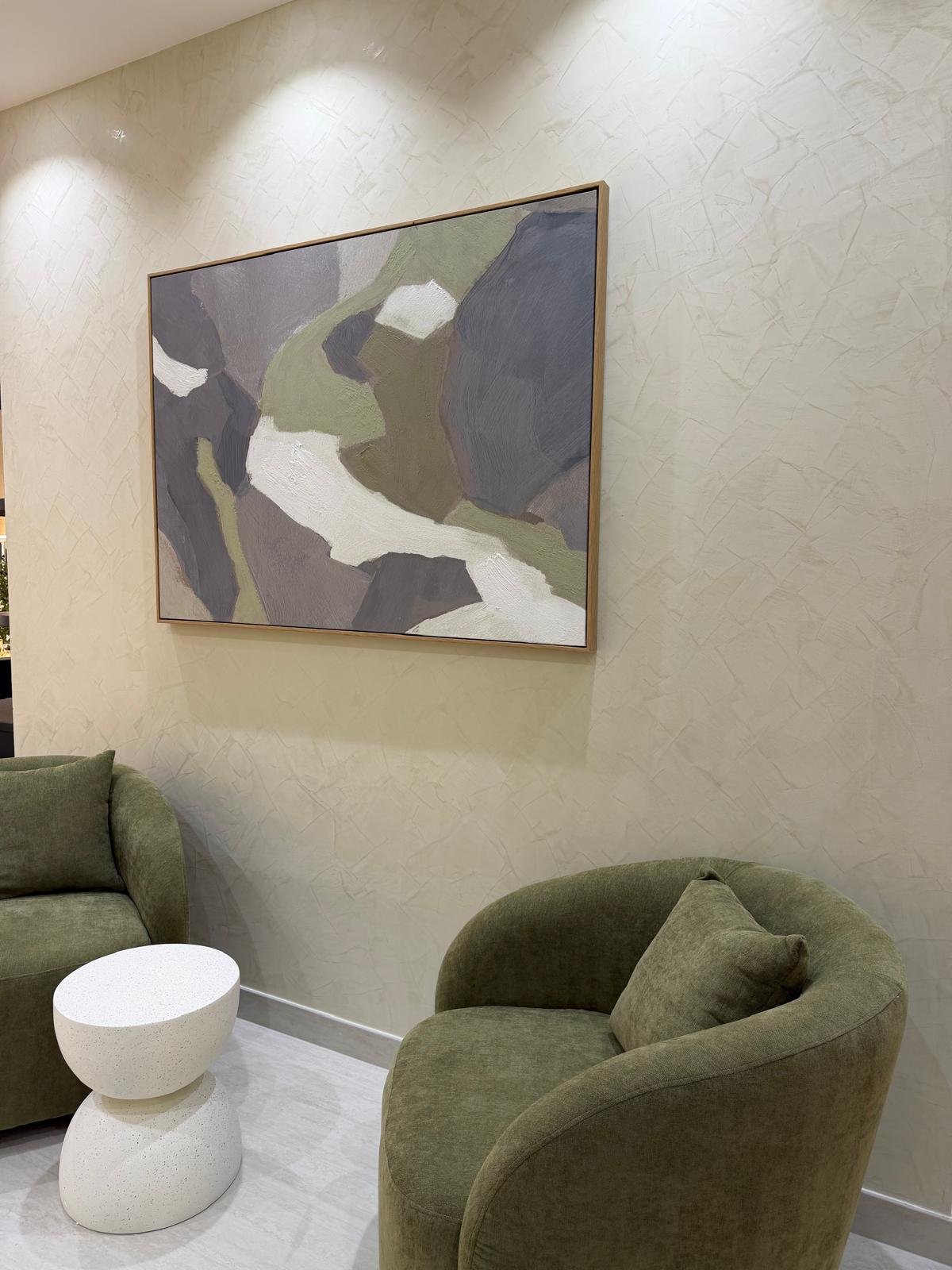 Waiting lounge at Brass Barber in Al Barsha, Dubai, with olive green armchairs, a white stone side table and a framed abstract painting on a plaster wall
